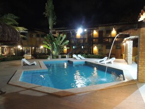 A nighttime swimming pool area with lounge chairs and a palm tree. - Marinas Pousada (Ilhéus)