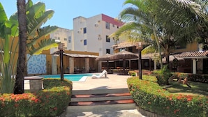 A pool area with a lounge chair, surrounded by tropical plants and a building with a tiled roof. - Marinas Pousada (Ilhéus)