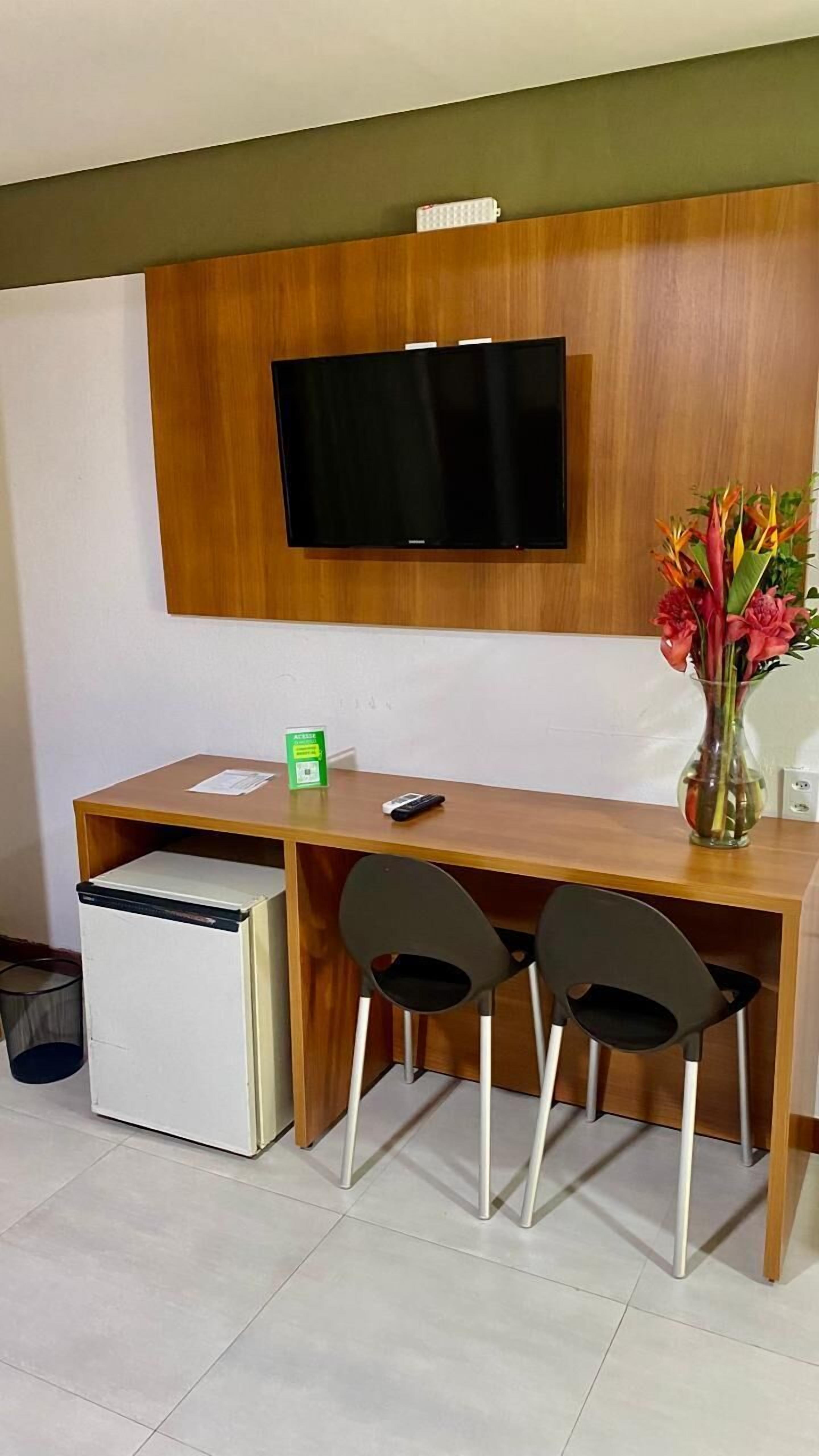 A compact room with a wooden desk, two chairs, a small refrigerator, a television mounted on the wall, and a vase with flowers.