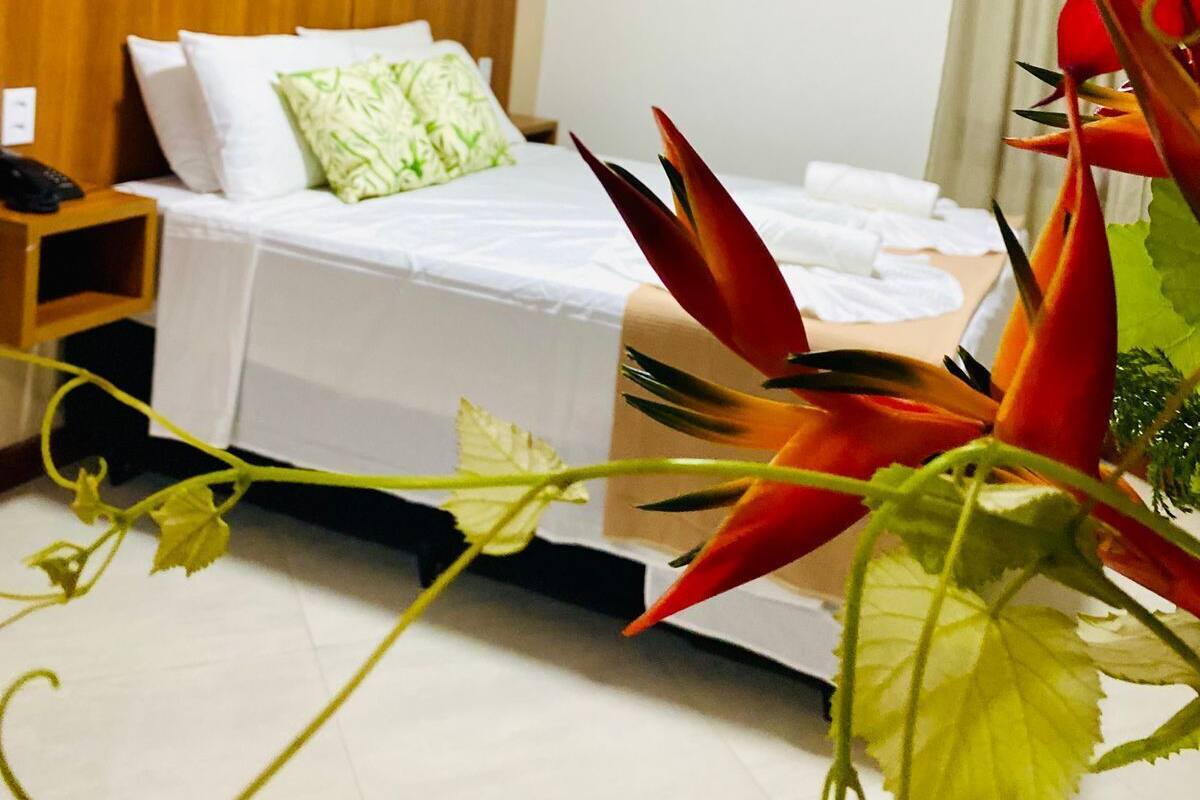 A hotel room with a bed, a nightstand, an air conditioning unit, and a colorful painting on the wall.