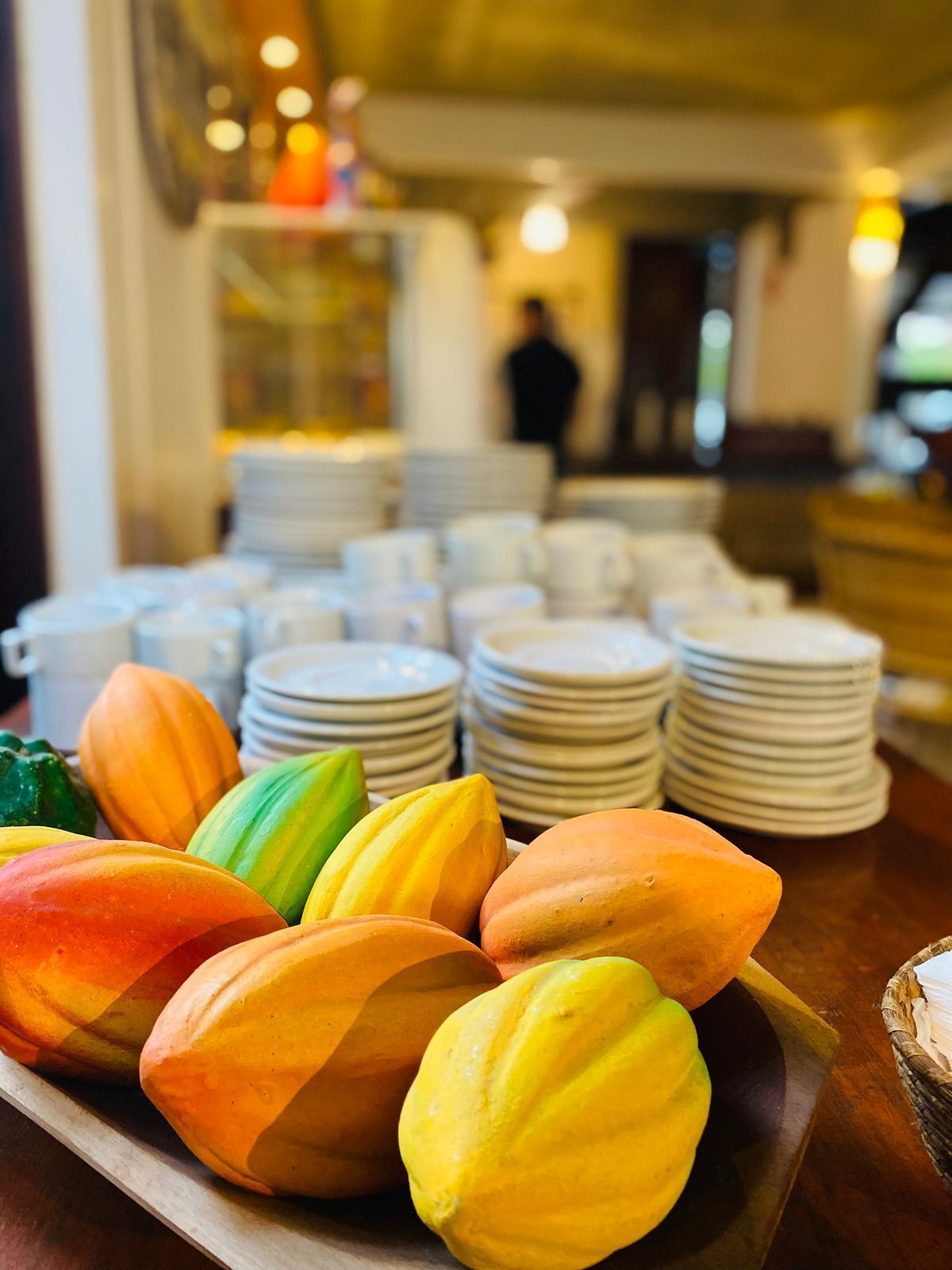 Uma bandeja de macarons coloridos sobre uma mesa de madeira, com pilhas de pratos brancos ao fundo.