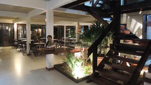 A spacious indoor area with multiple dining tables and chairs, a central planter with plants, and a staircase with a wooden railing. - Marinas Pousada (Ilhéus)