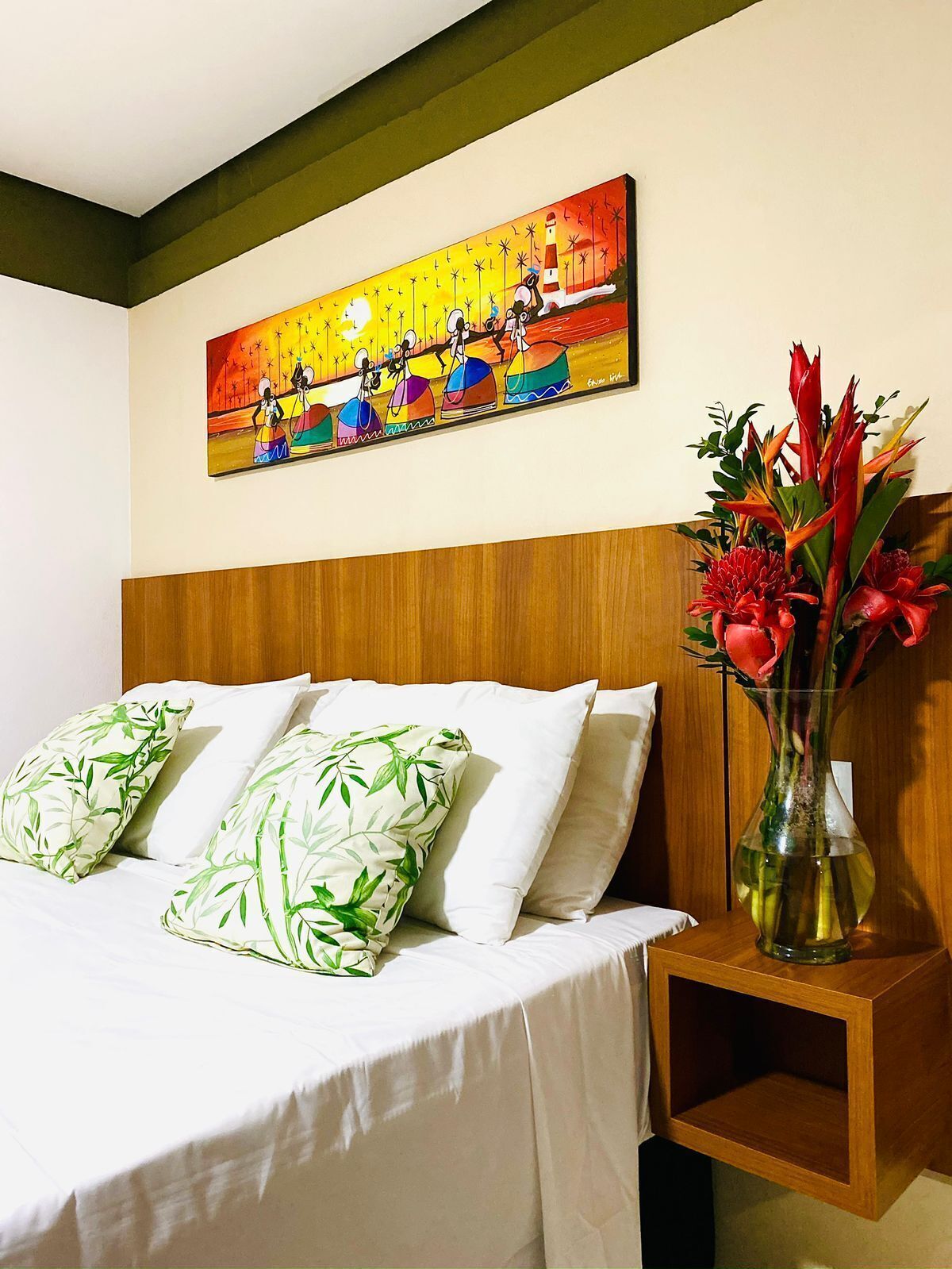 A hotel room with a bed, pillows, a nightstand, a vase with flowers, and a painting on the wall.