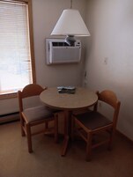 Una mesita redonda pequeña con dos sillas de madera, una lámpara de mesa y un equipo de aire acondicionado instalado en la pared.