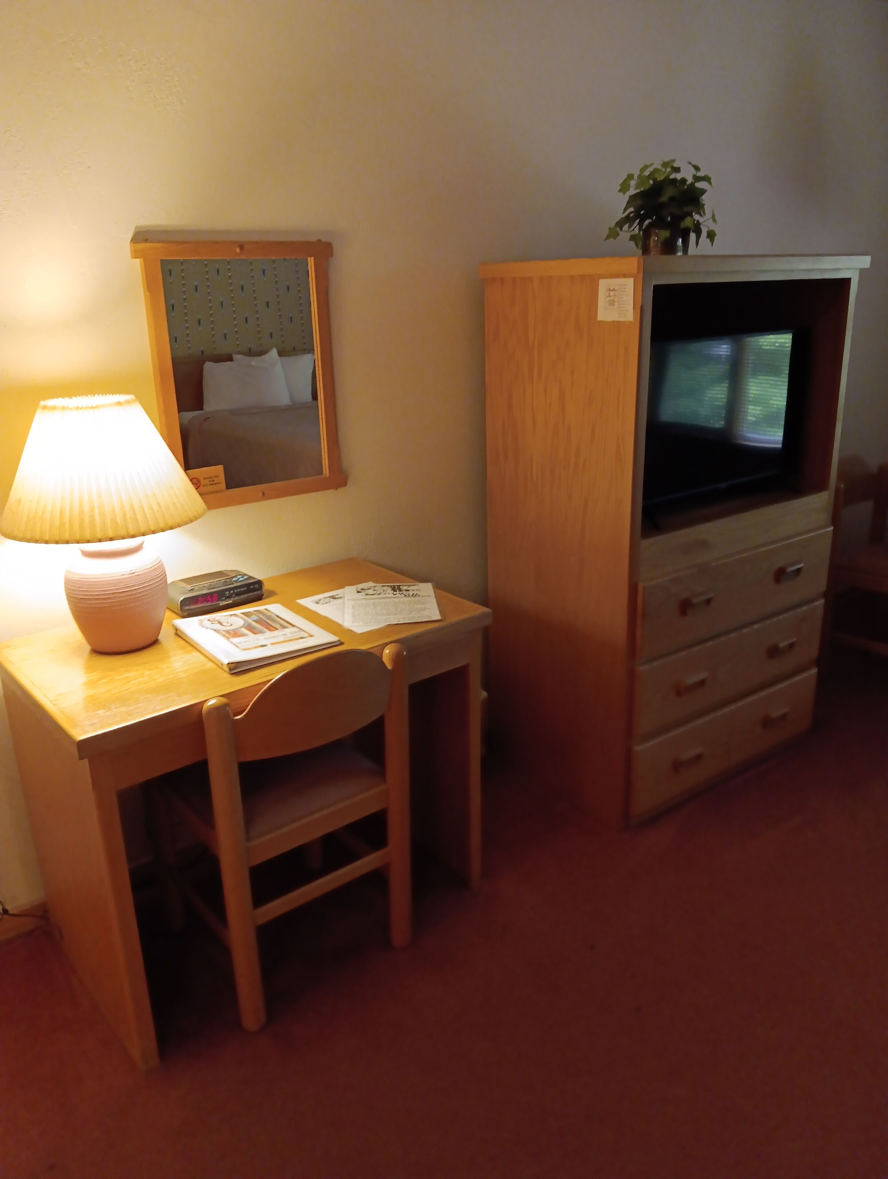 A small hotel room with a wooden desk, a lamp, a television, and a mirror.