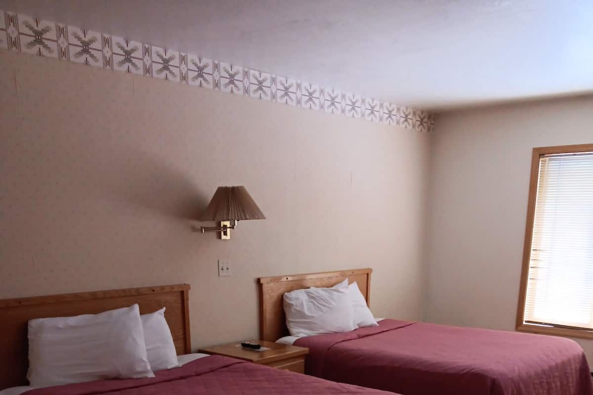 A hotel room with two beds, a nightstand, a lamp, and a window with blinds.