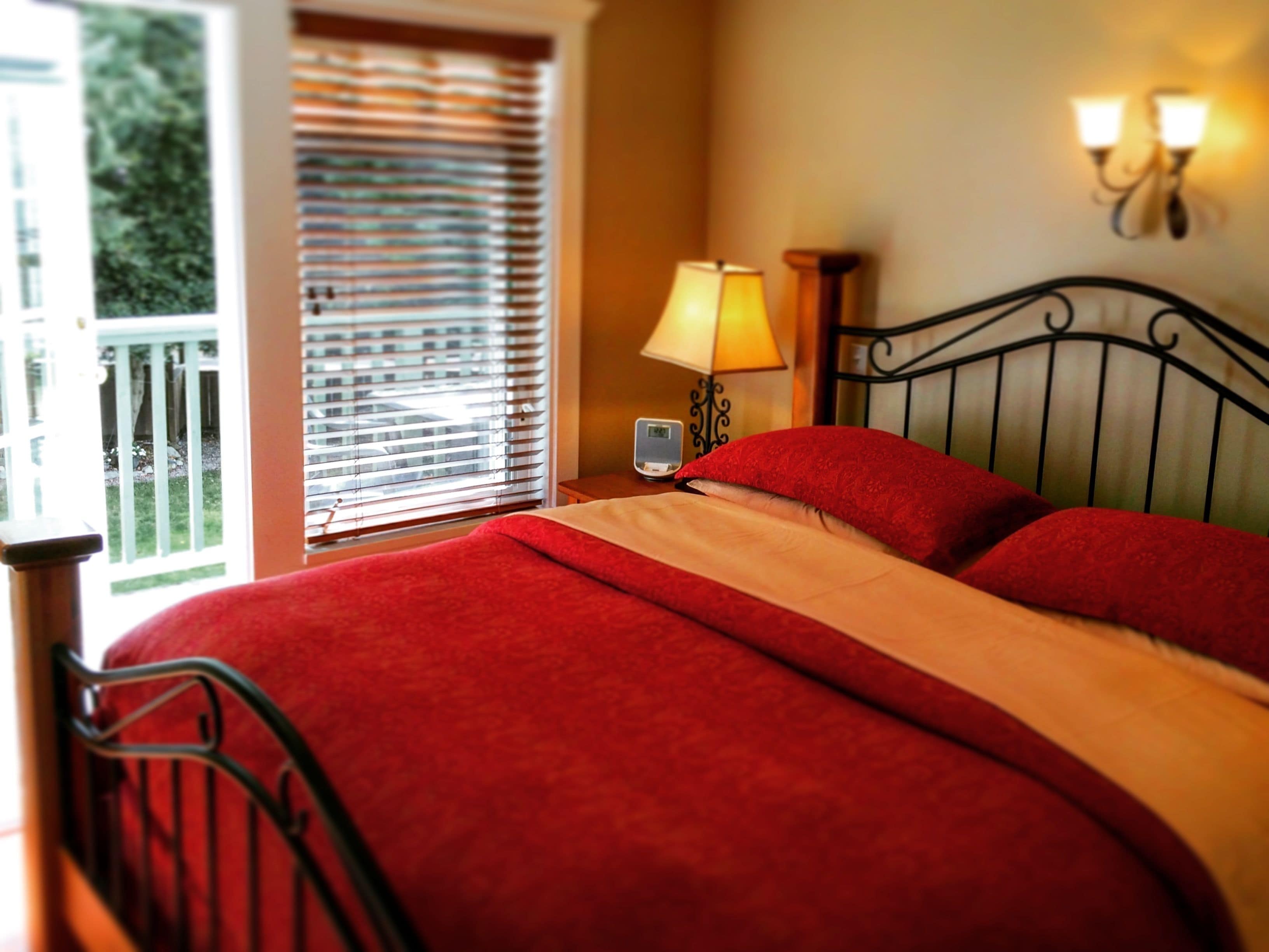 Una cama con ropa de cama roja, una mesita de noche con lámpara y una ventana con persianas.