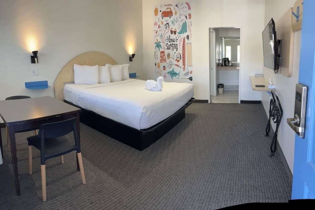A hotel room with a bed, a desk, a chair, a wall with a surfing-themed decoration, and a TV mounted on the wall.