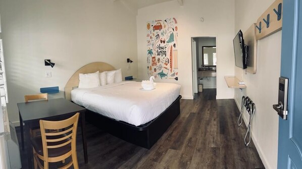 A bedroom with a bed, a desk, a chair, a TV, and a wall with a surfing-themed decoration. - Ocean Palms Motel (Pismo Beach)