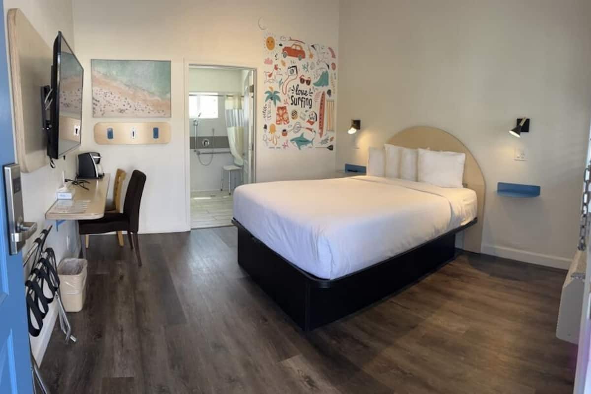 A bedroom with a bed, a desk, a chair, a TV, and a bathroom visible through an open door.