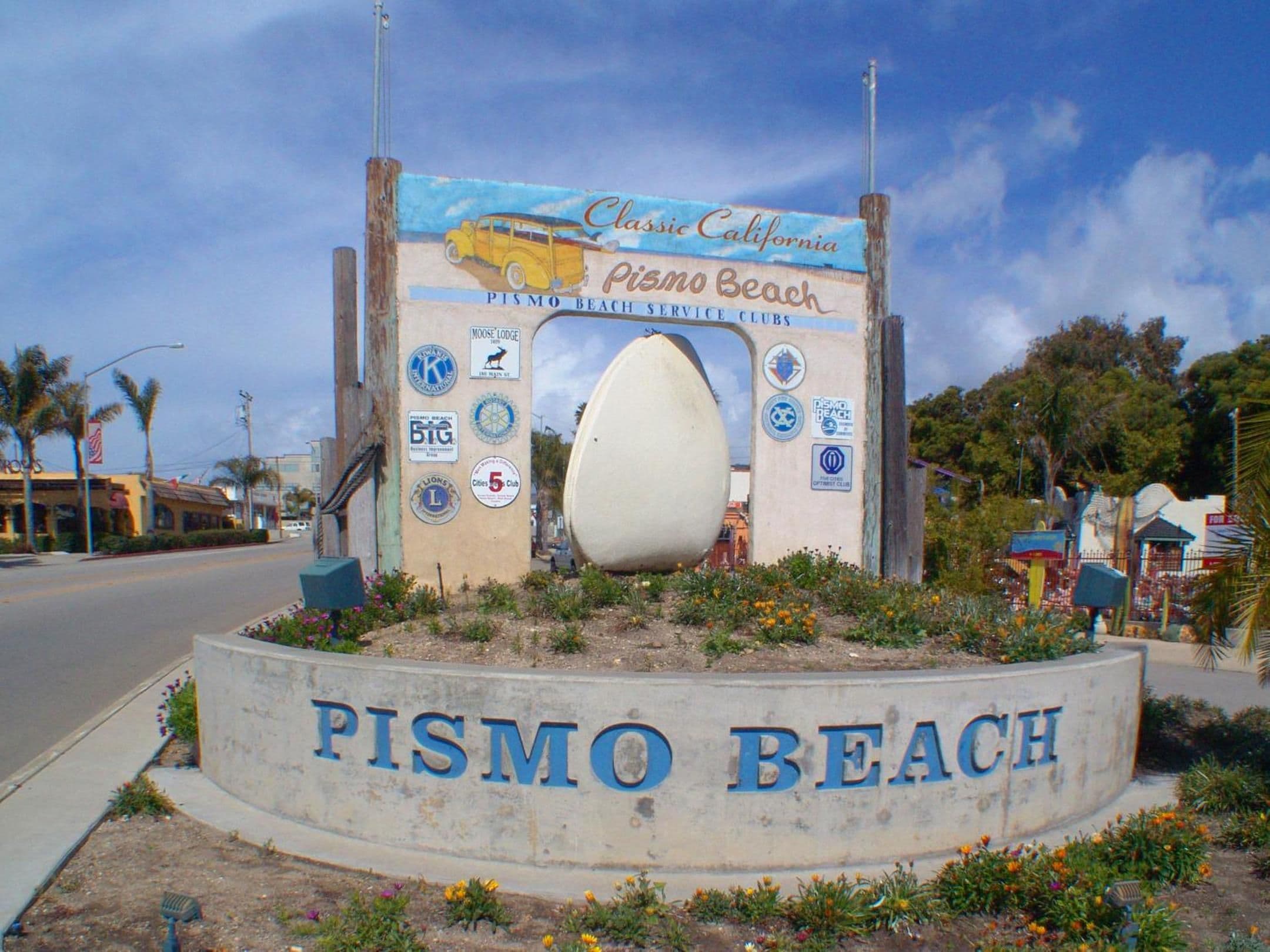 A sign for Pismo Beach with a large surfboard and various logos.