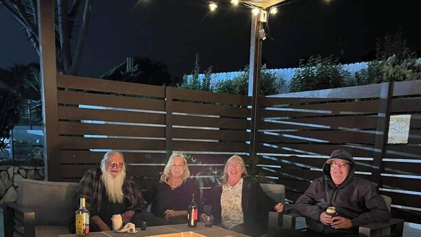 A stone fire pit with three people seated around it, under a covered area with string lights. - Ocean Palms Motel (Pismo Beach)
