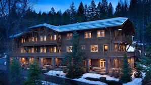A large, multi-story lodge with a prominent balcony and a stone foundation, surrounded by a snowy landscape and dense forest. - Belton Chalet (West Glacier)