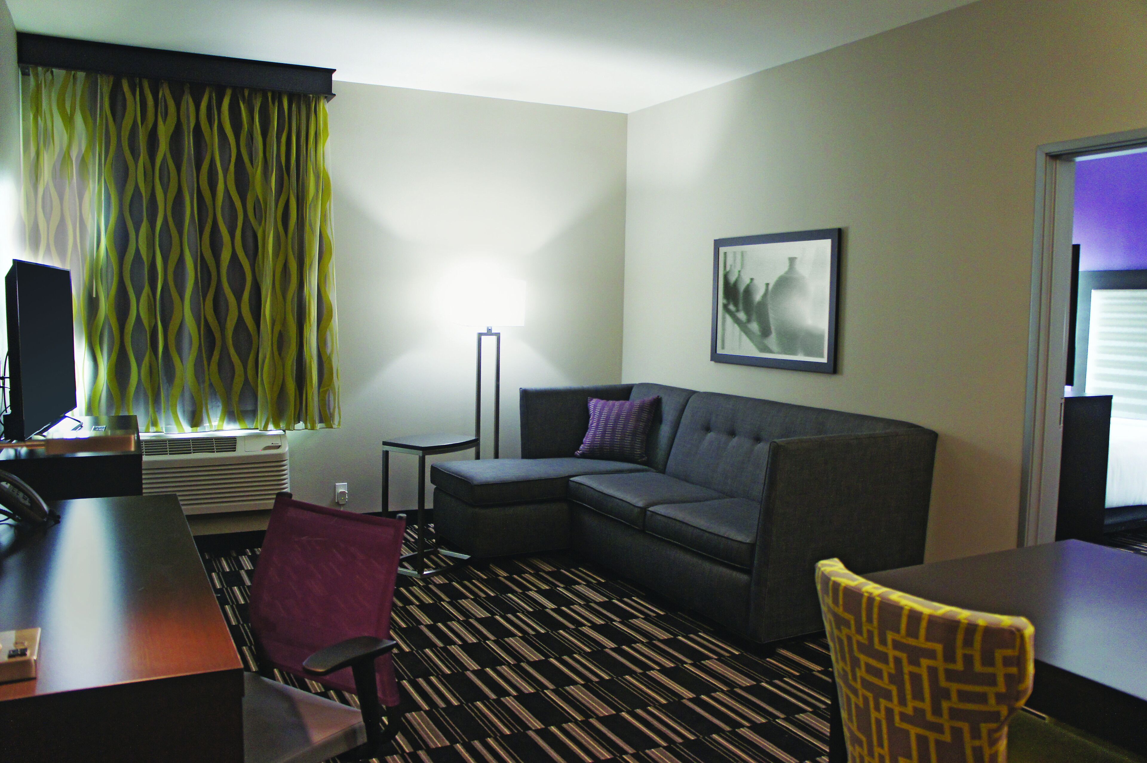 A hotel room with a sectional sofa, a desk with a chair, a standing lamp, and a framed picture on the wall.