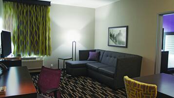 A hotel room with a sectional sofa, a desk with a chair, a standing lamp, and a framed picture on the wall.