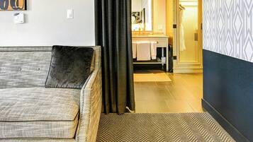 A hotel room with a sofa, a patterned rug, a shower area, and a bathroom with a towel rack.