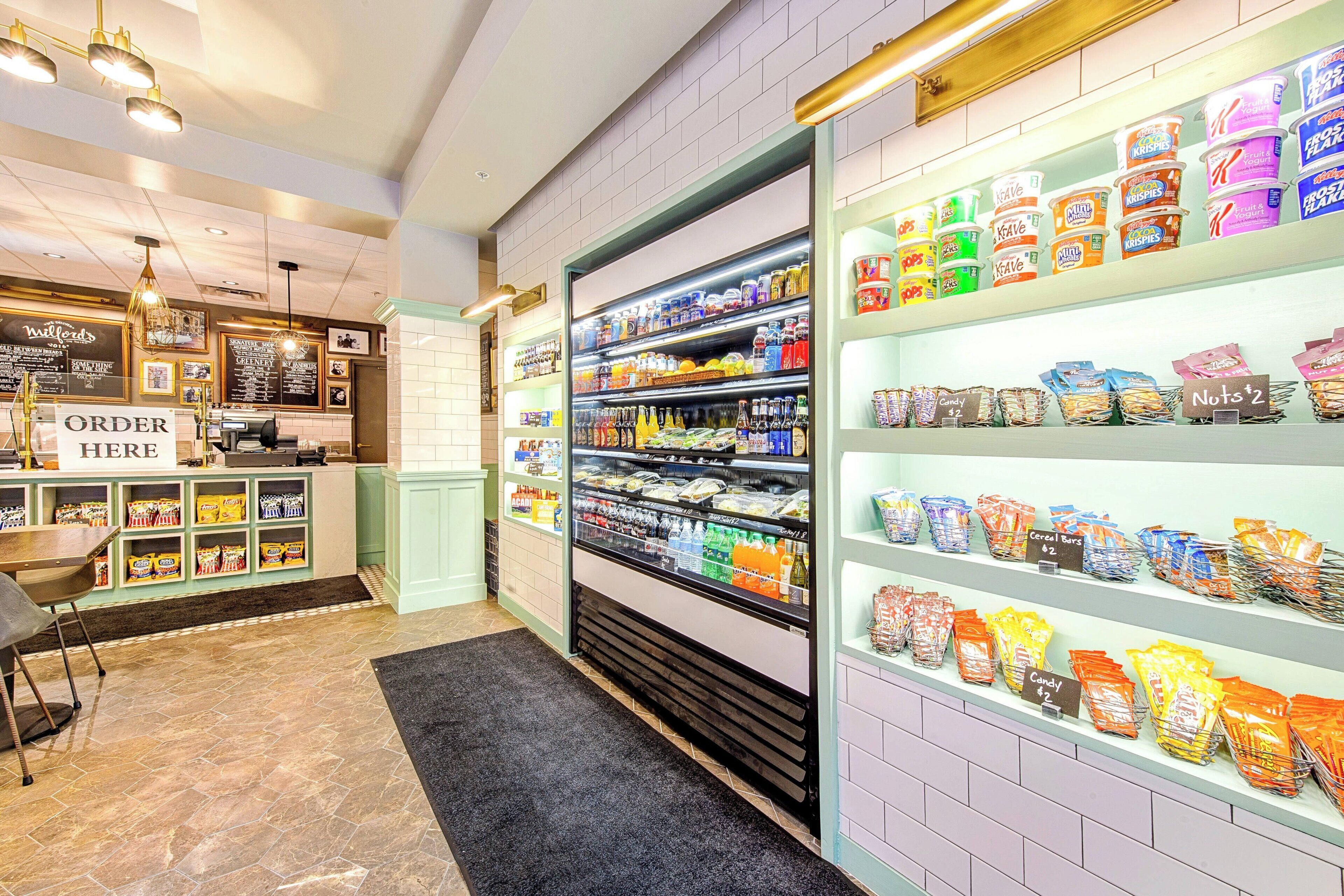 Um bar de lanches com diversos produtos embalados, bebidas, um balcão com a placa "ORDER HERE" e uma área de estar com mesa e cadeiras.