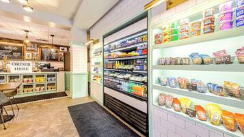 Um bar de lanches com diversos produtos embalados, bebidas, um balcão com a placa "ORDER HERE" e uma área de estar com mesa e cadeiras.