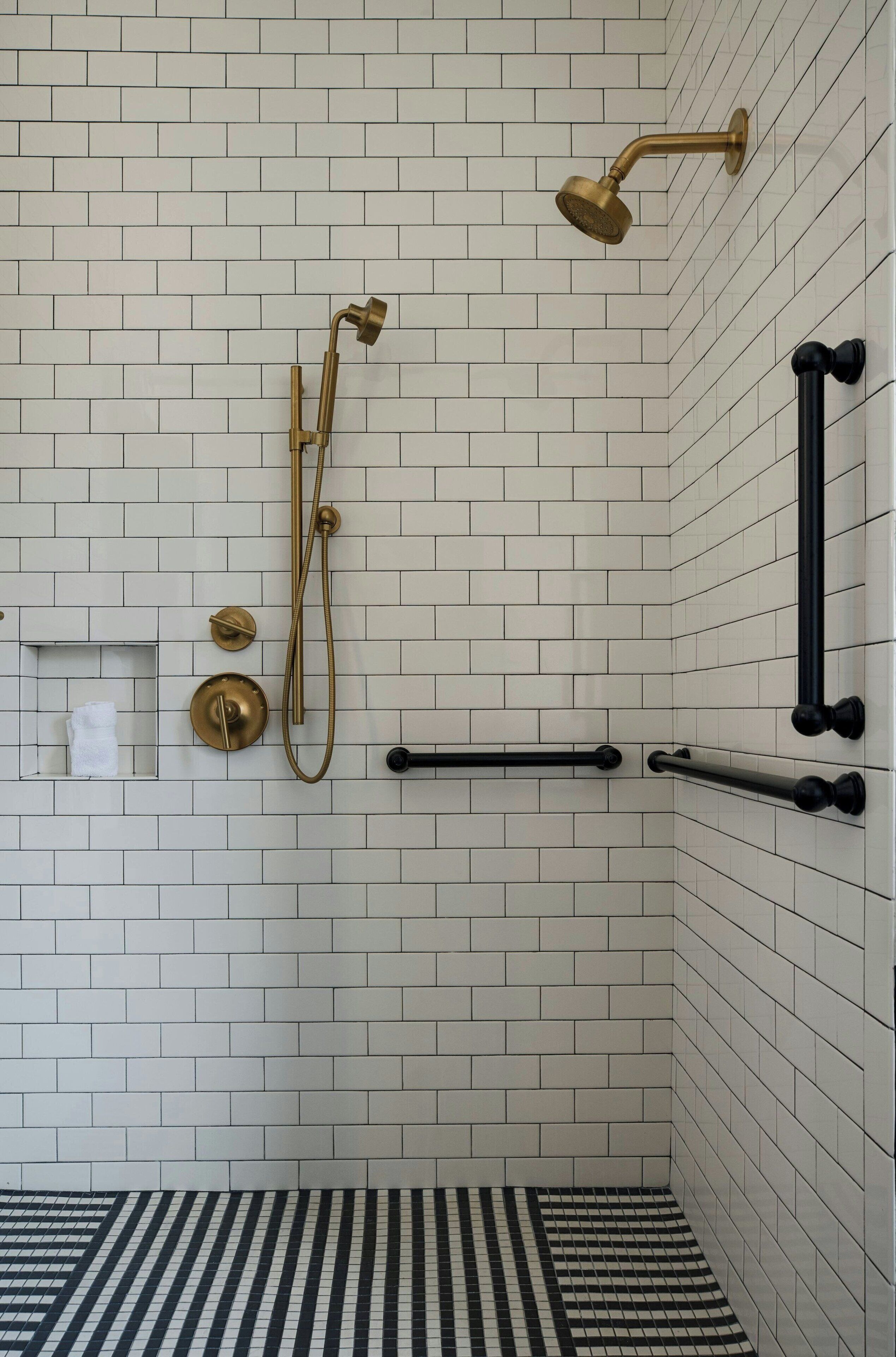 A bathroom with white tiled walls, a black and white striped floor, and a gold shower fixture.