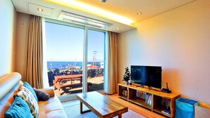 A living room with a sofa, a coffee table, a TV on a wooden stand, and a view of the ocean through large windows. - JejuSorang (Jeju City)