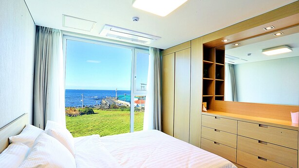 A hotel room with a large bed, a wooden dresser, and a view of the ocean. - JejuSorang (Jeju City)