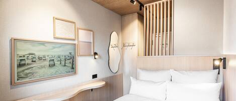 A modern hotel room with a wooden ceiling, a bed with white linens, a bedside table, a wall-mounted mirror, and artwork on the wall.