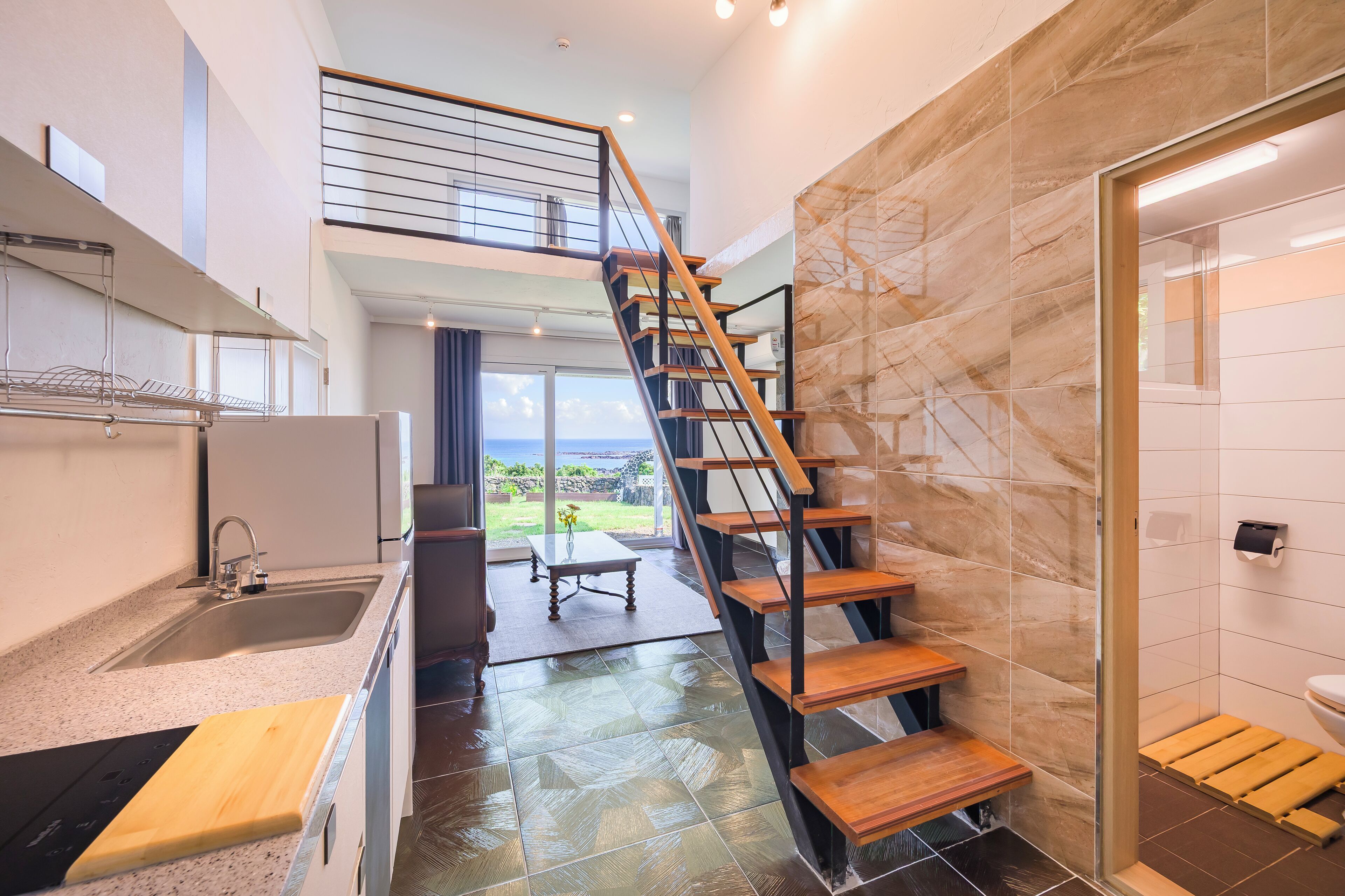 A modern kitchen with a large island, a wooden staircase, and a view of the outdoors.
