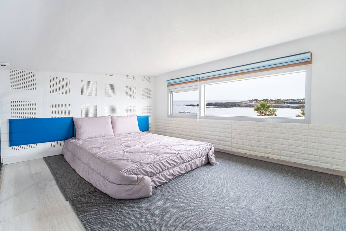 A bedroom with a bed, a window with a view of the sea, and a white brick wall.