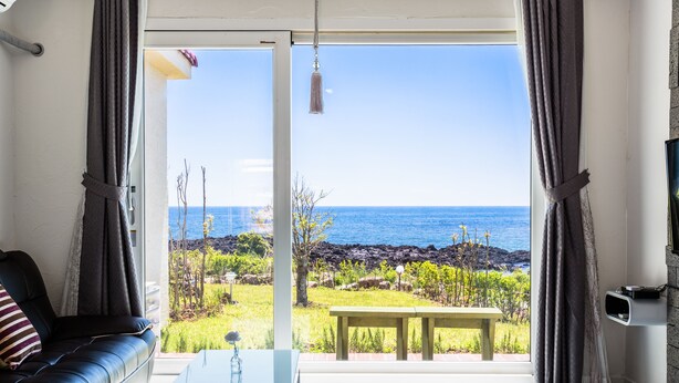 A living room with a black leather sofa, a glass coffee table, and a view of the ocean through large windows. - Jeju Turning Point (Seogwipo)