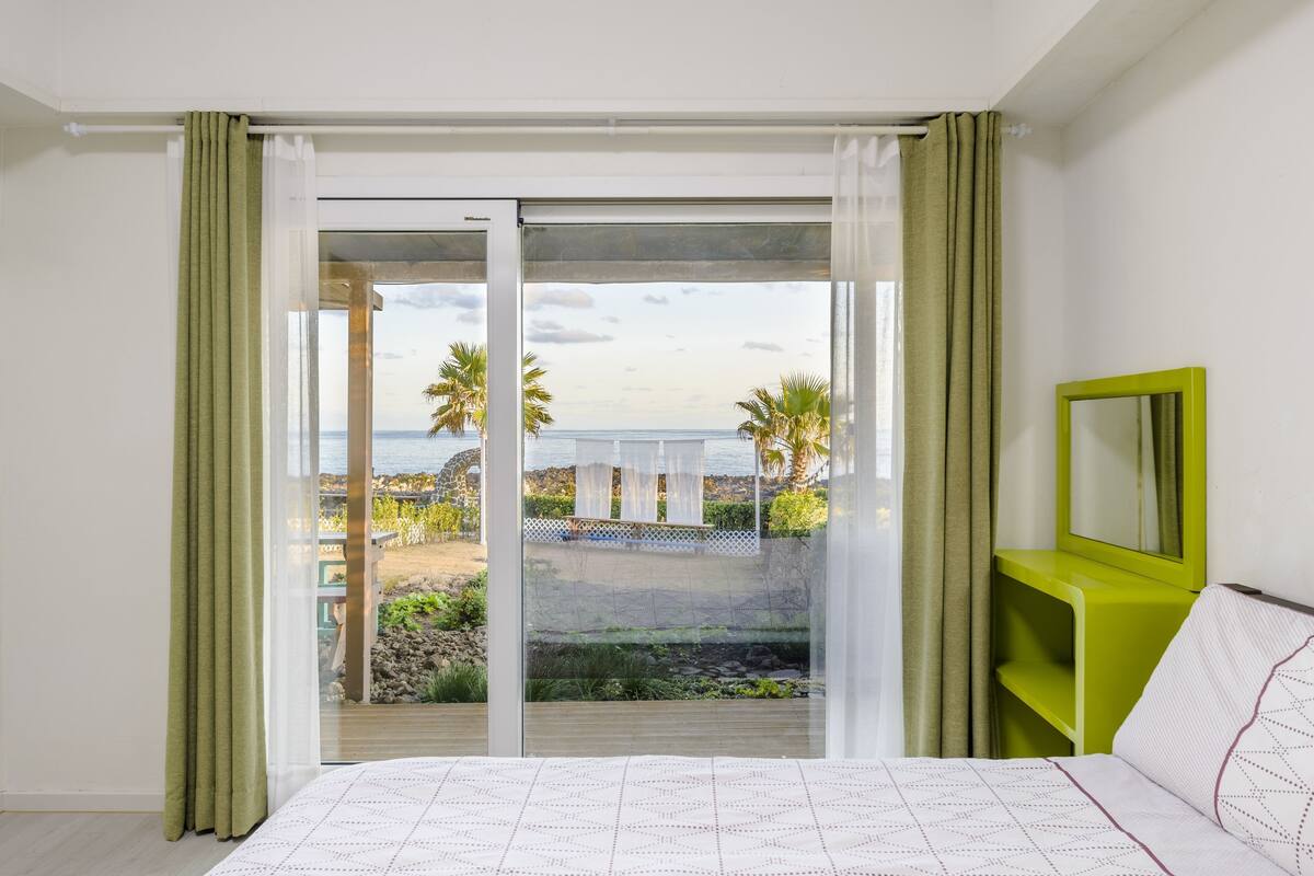A bedroom with a bed, a green nightstand, a mirror, and a view of the beach and palm trees through a sliding glass door.