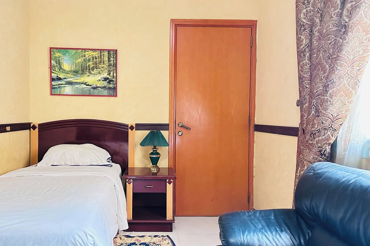 A bedroom with a single bed, a nightstand with a lamp, a door, and a blue chair.