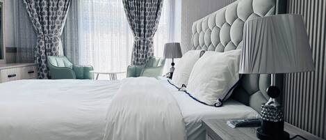 A modern hotel room with a large bed, a nightstand with a lamp, a chair, and a view of the city through sheer curtains.