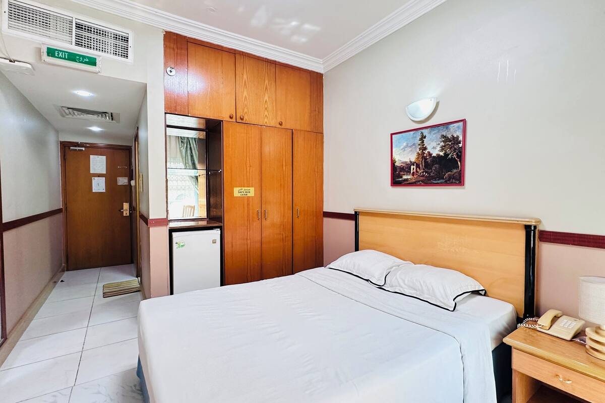 A hotel room with a bed, a desk with a telephone, a small refrigerator, a wardrobe, and an exit sign.