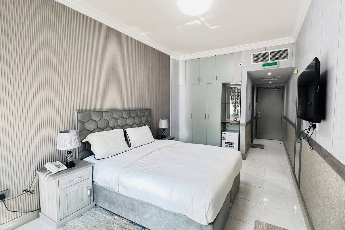 A modern hotel room with a large bed, a bedside table with a lamp, a wardrobe, a television mounted on the wall, and an exit sign.