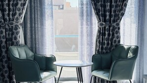 A room with two green chairs and a small round table, curtains with a geometric pattern, and a view of buildings through the window. - Palette San Marino Hotel (Dubai)
