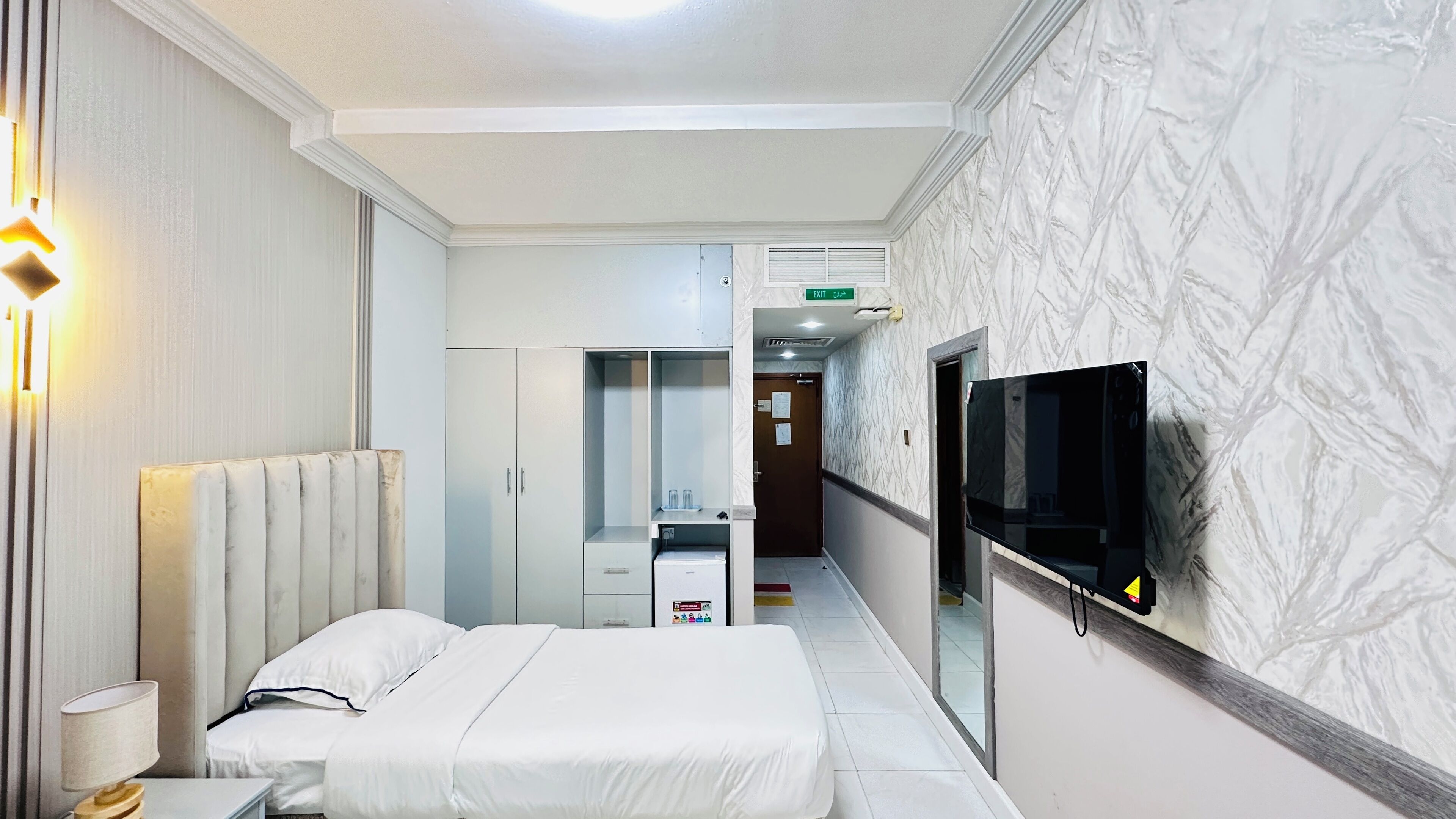 Habitación de hotel con cama, mesita de noche, lámpara, armario, espejo y televisor instalado en la pared.