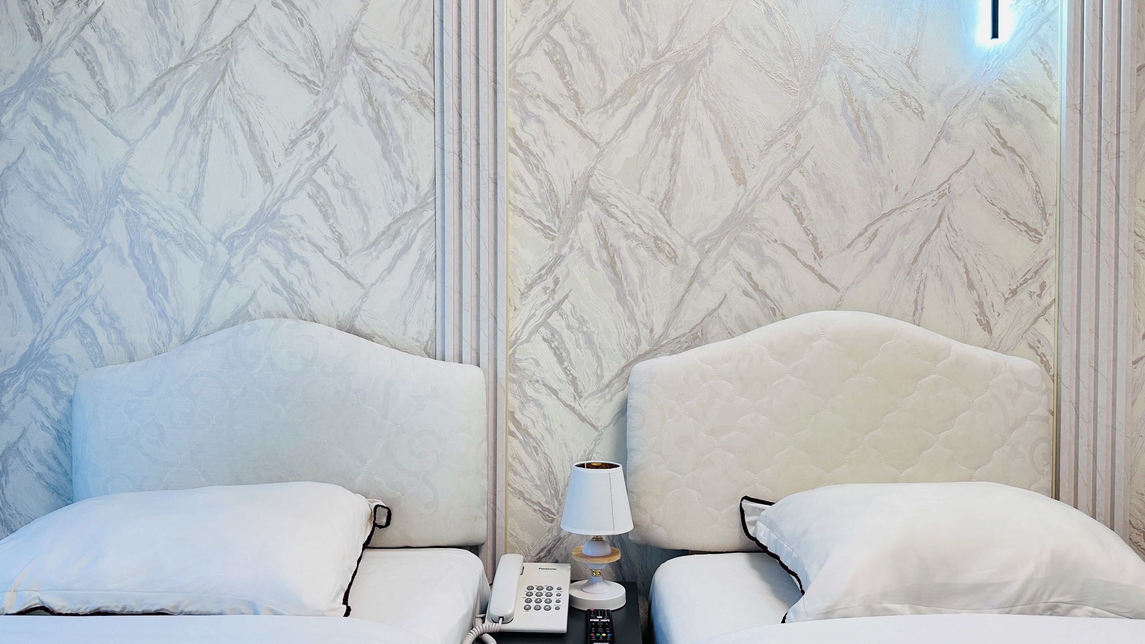 A hotel room with a marble-patterned wall, a bed with white bedding, a bedside table with a lamp, a phone, and a remote control.