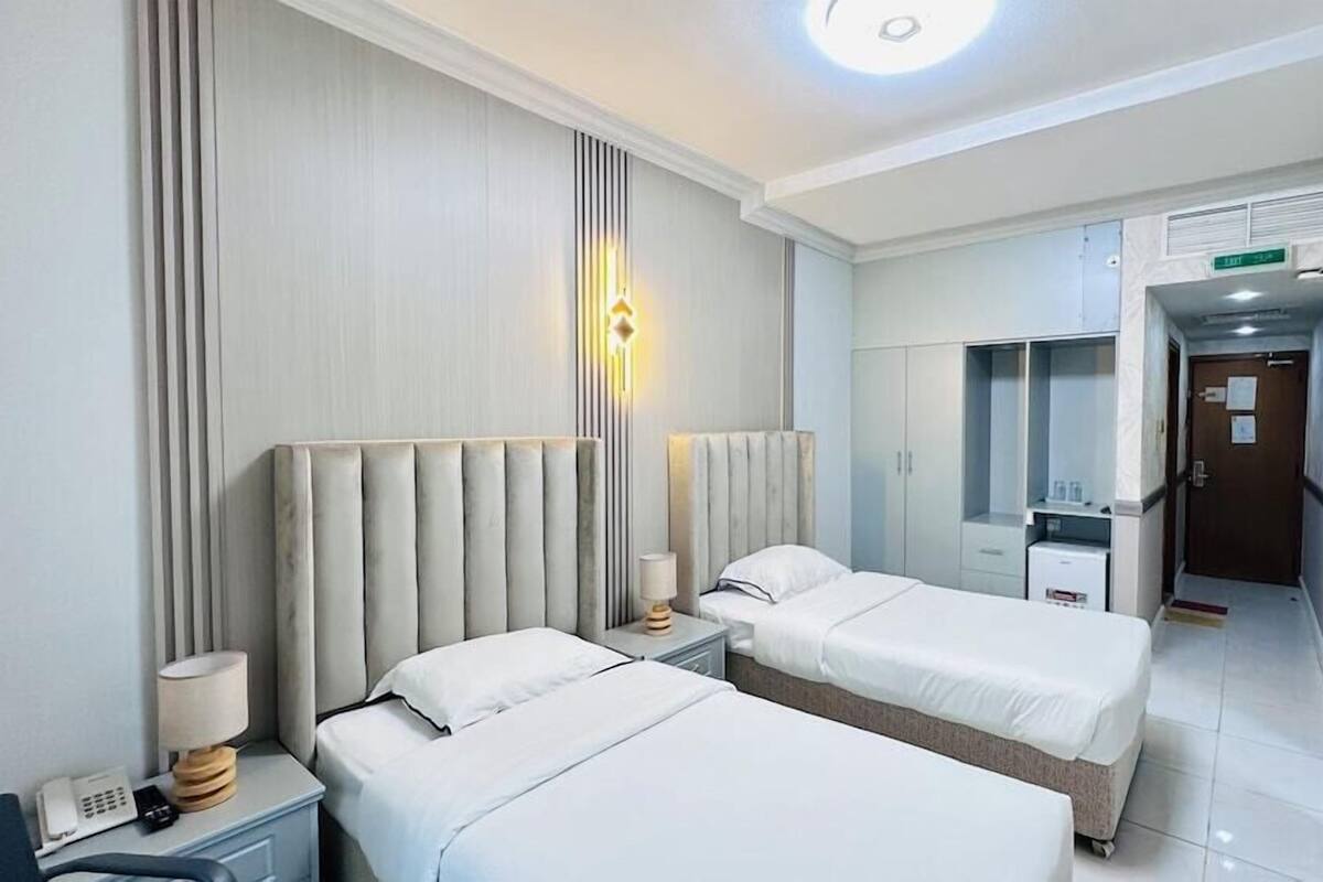 A hotel room with two beds, a bedside table with a lamp, a telephone, a wardrobe, and a television mounted on the wall.
