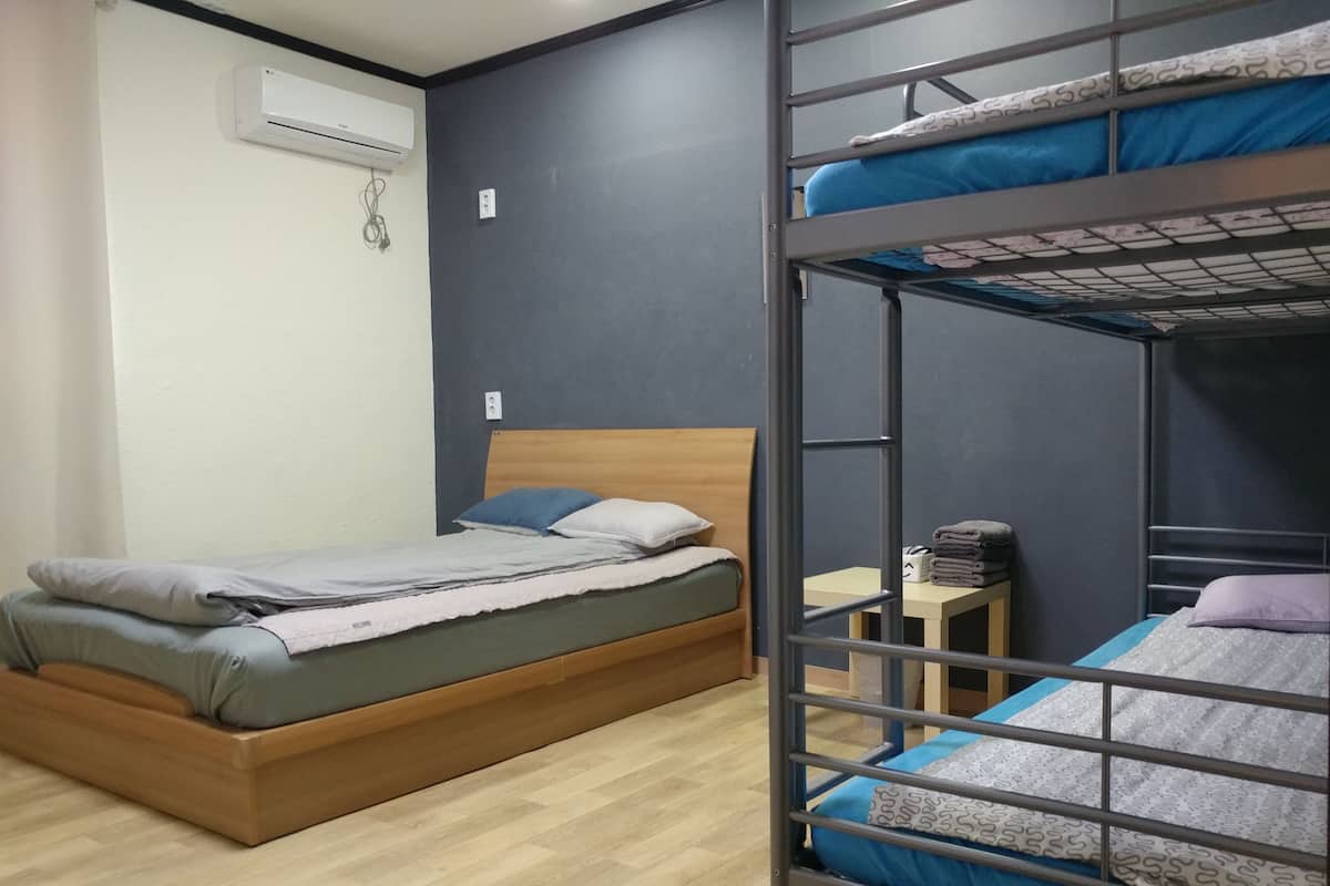 A dormitory room with bunk beds, a single bed, wooden flooring, an air conditioning unit, and a small table with a towel.