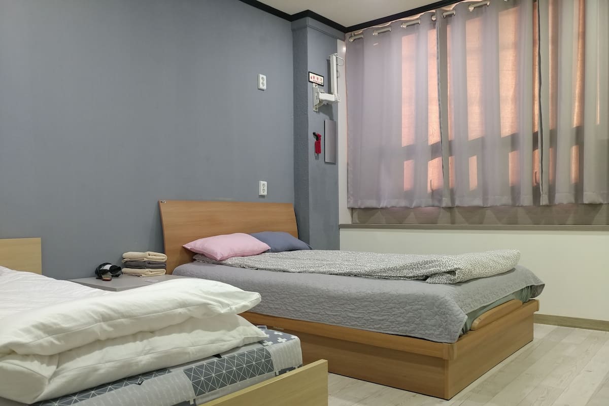 A bedroom with two beds, a window with sheer curtains, and a wall-mounted light fixture.