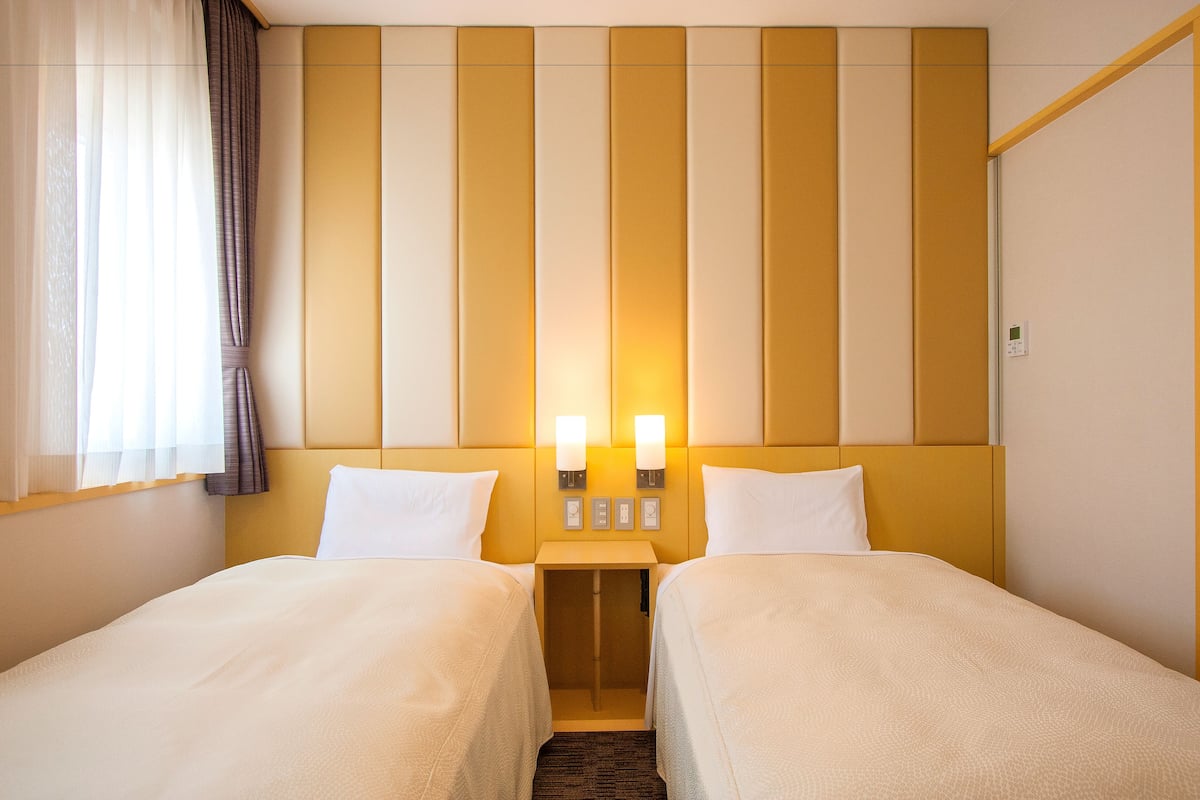 A hotel room with two beds, a headboard with vertical panels, two wall-mounted lights, and a small table between the beds.