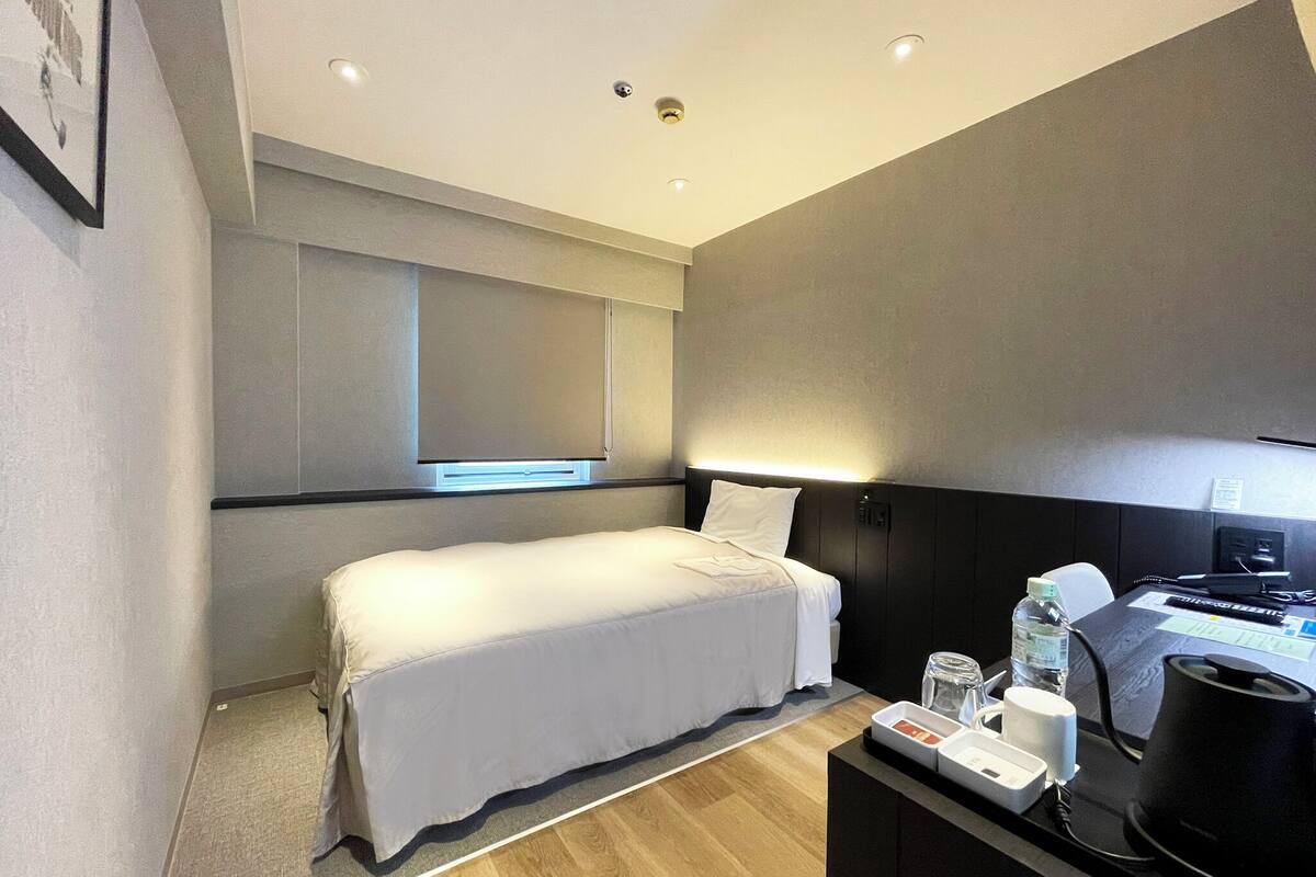 A modern hotel room with a single bed, a desk with a kettle, and a small table with a bottle and glasses.