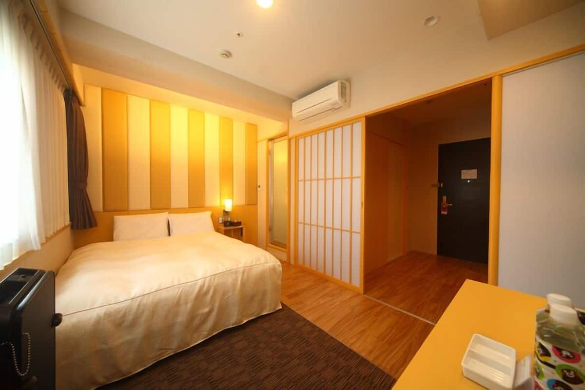 A hotel room with a bed, bedside table, lamp, air conditioning unit, and a sliding door leading to another room.