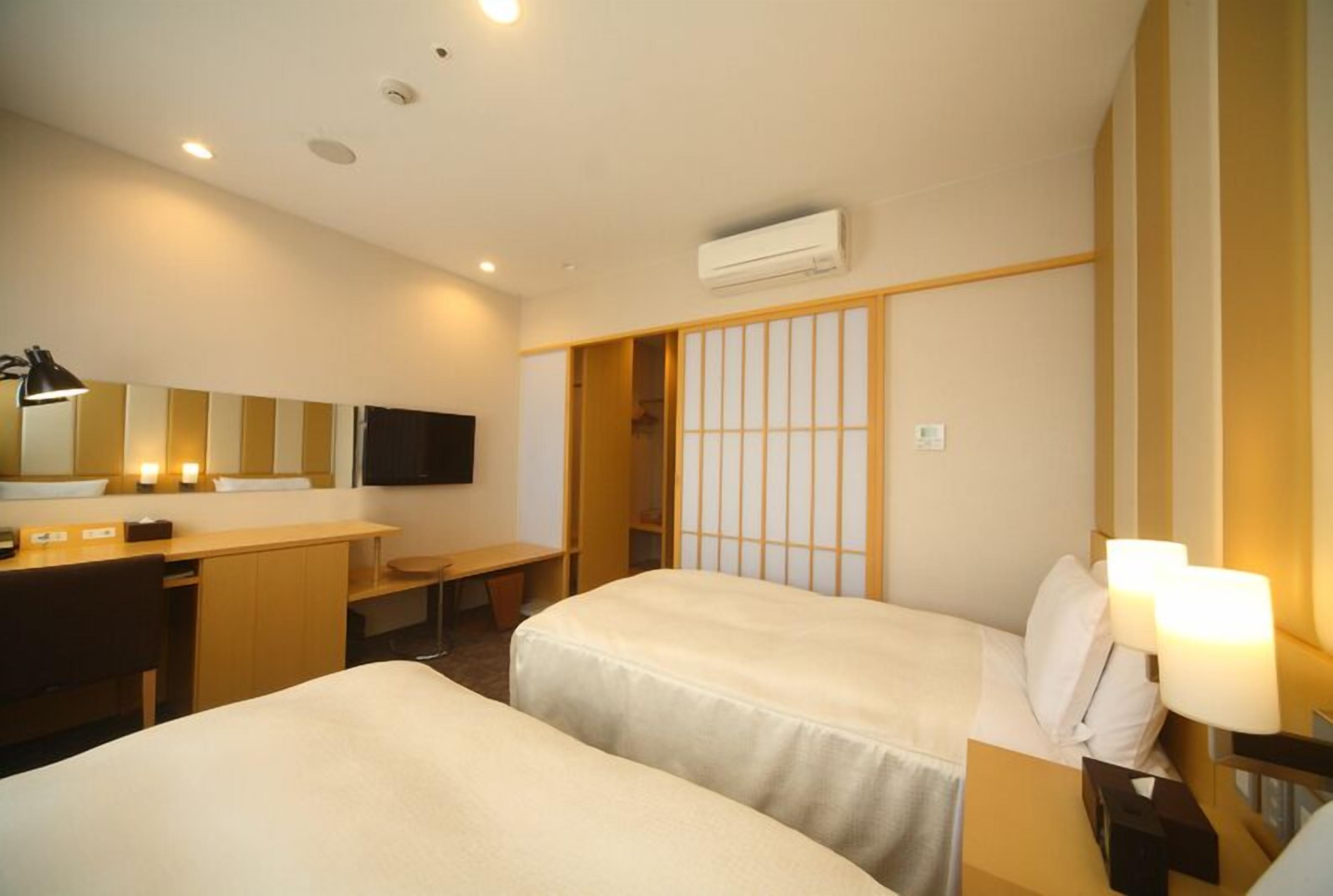 A hotel room with two beds, a desk, a chair, a TV, and a sliding door.