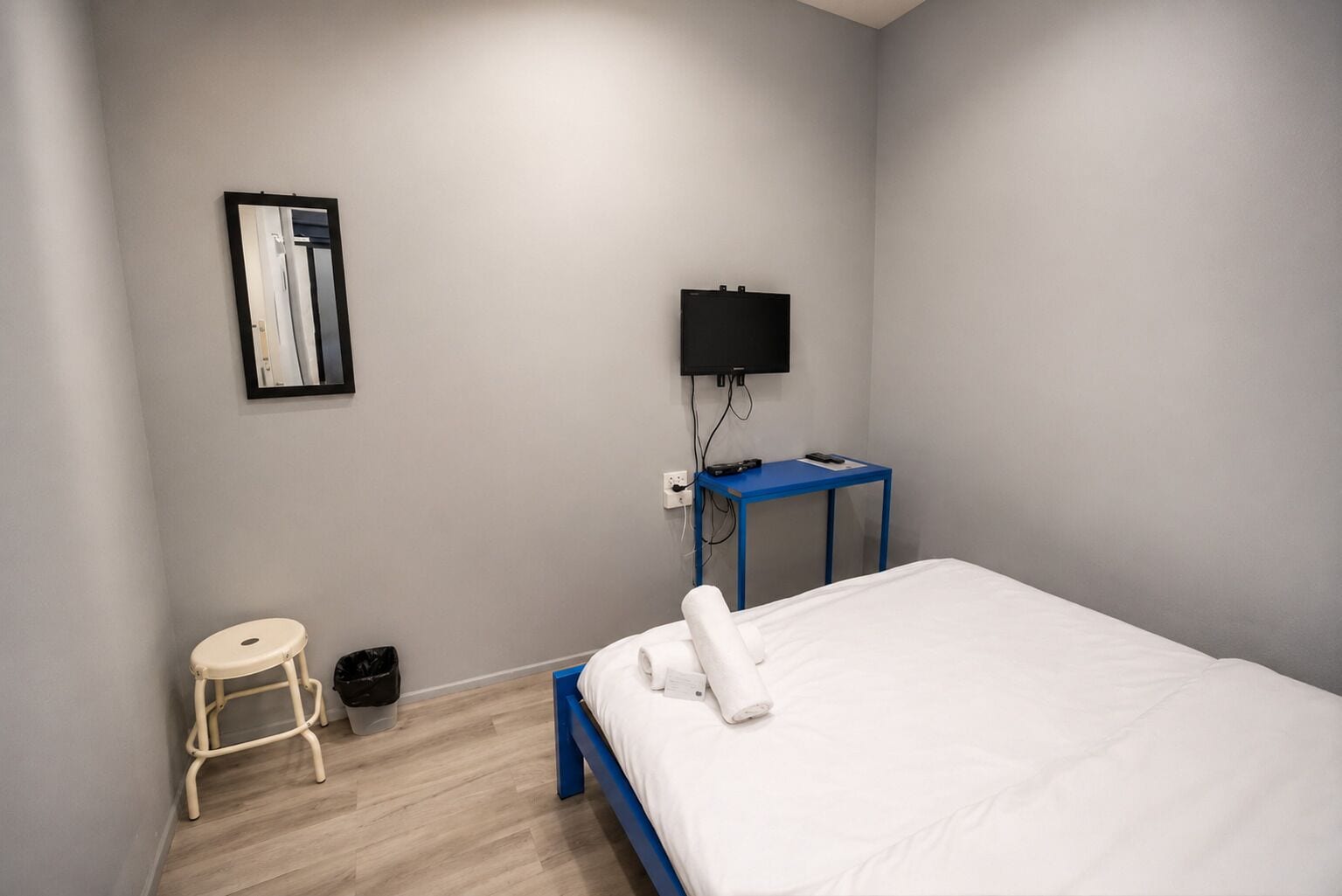 Camera d'albergo con un letto, uno sgabello, un cestino, una scrivania con un televisore e uno specchio fissato alla parete.
