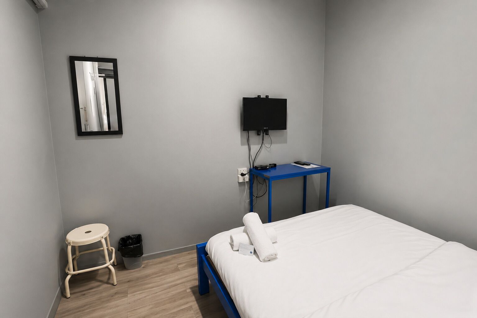 A hotel room with a bed, a small stool, a trash bin, a blue table, and a TV mounted on the wall.