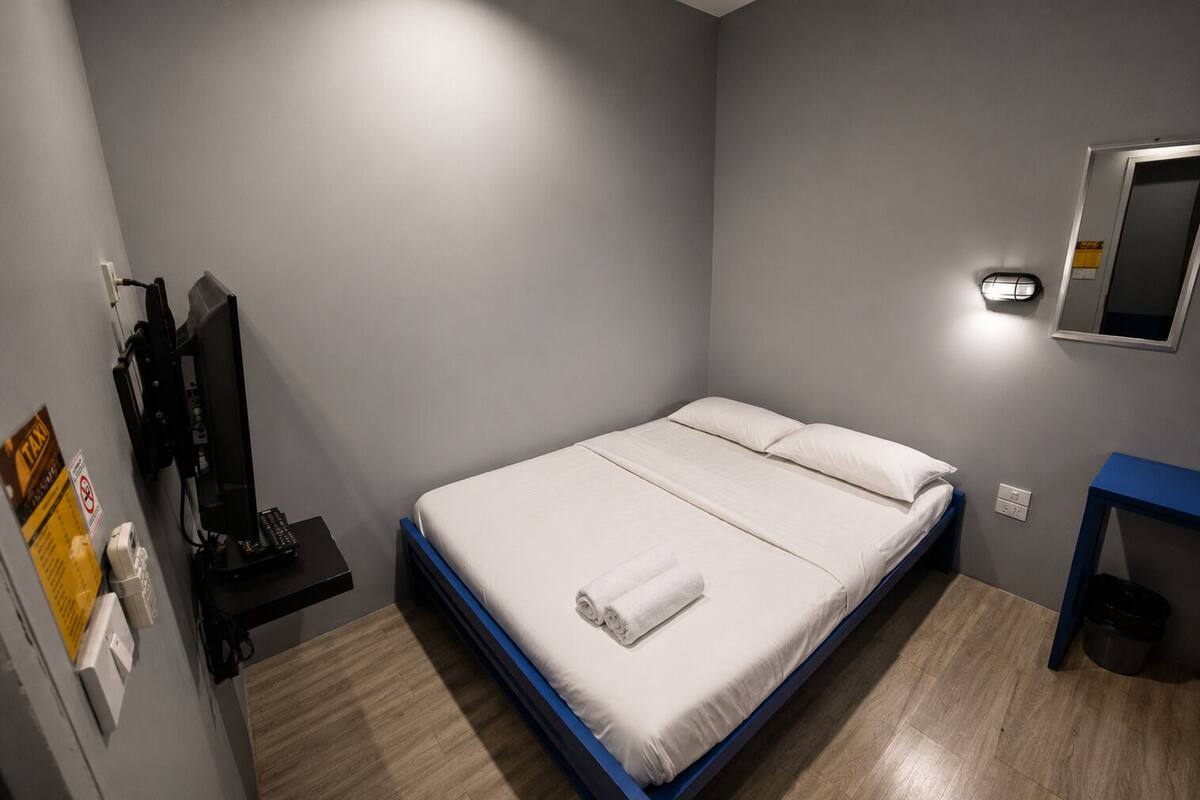 A single bed with a white mattress and blue frame, a small bedside table, a wall-mounted TV, and a mirror.