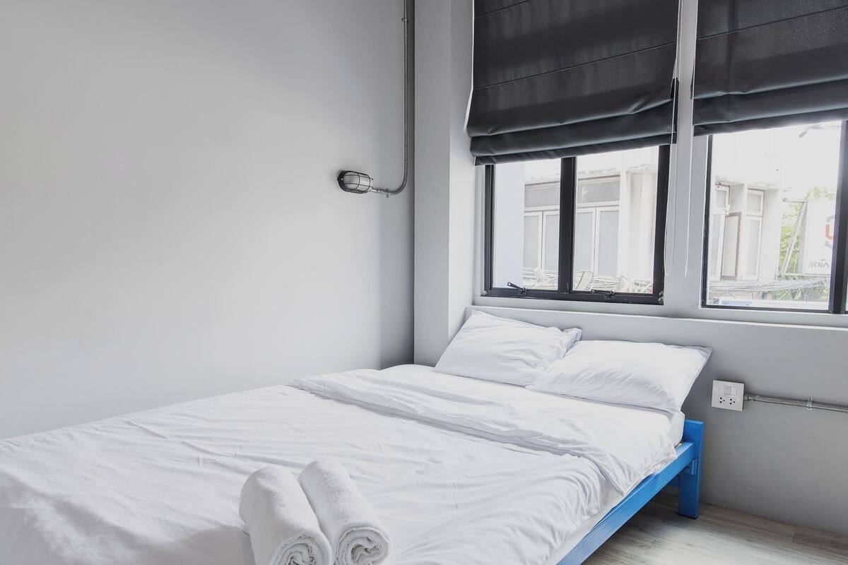 A single bed with white bedding, a window with black curtains, and a visible electrical outlet on the wall.