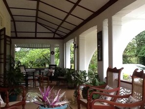 A covered patio area with a table and chairs, surrounded by potted plants and columns.