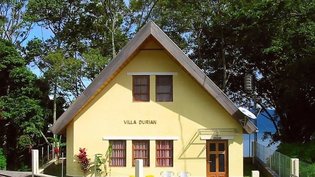 A yellow villa named "Villa Durián" with a triangular roof, two windows, a door, and a small outdoor seating area with chairs.